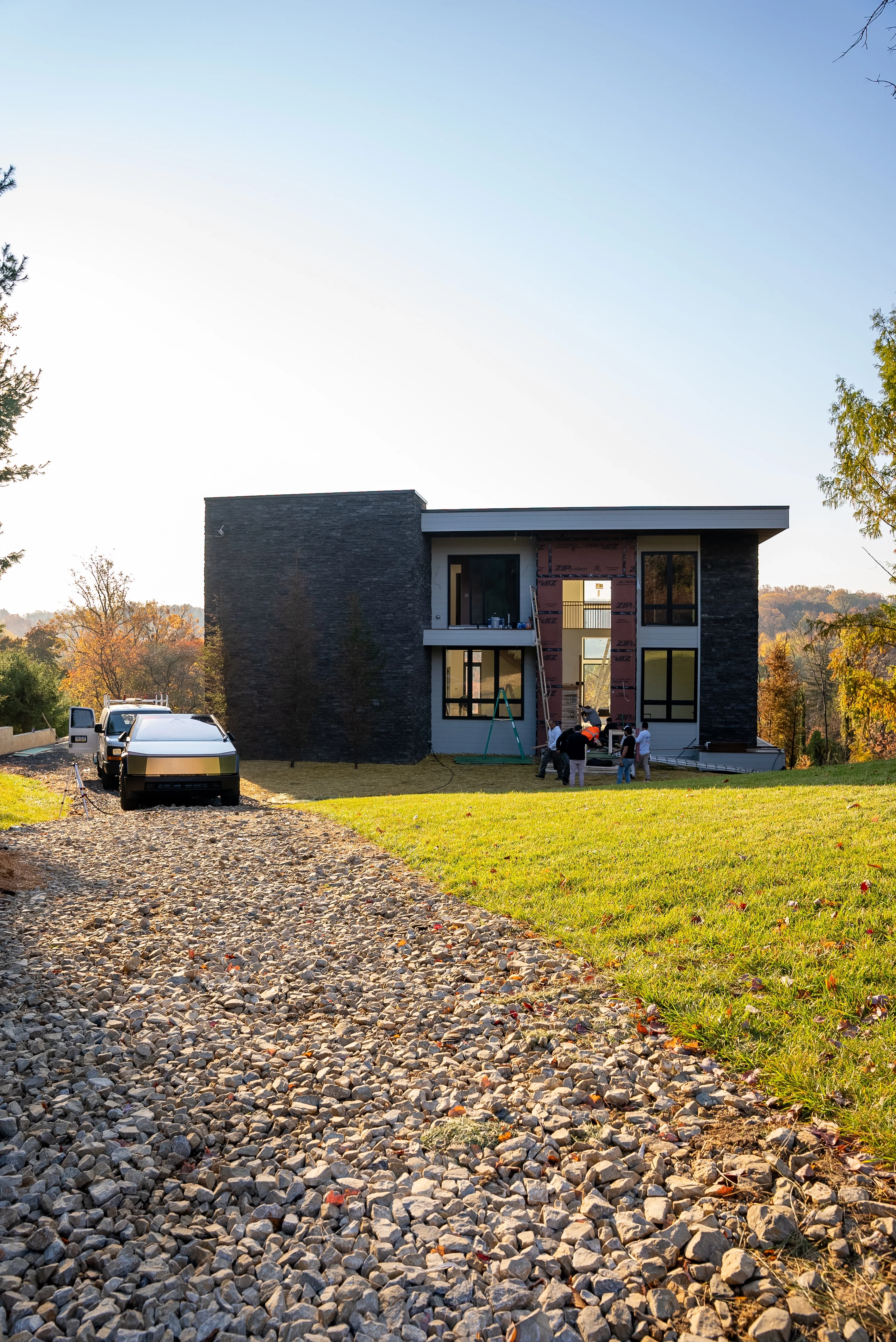 A modern two-story home under construction, featuring large glass windows and a stone façade, with a gravel driveway leading up to the property, surrounded by an expansive lawn and scenic views.