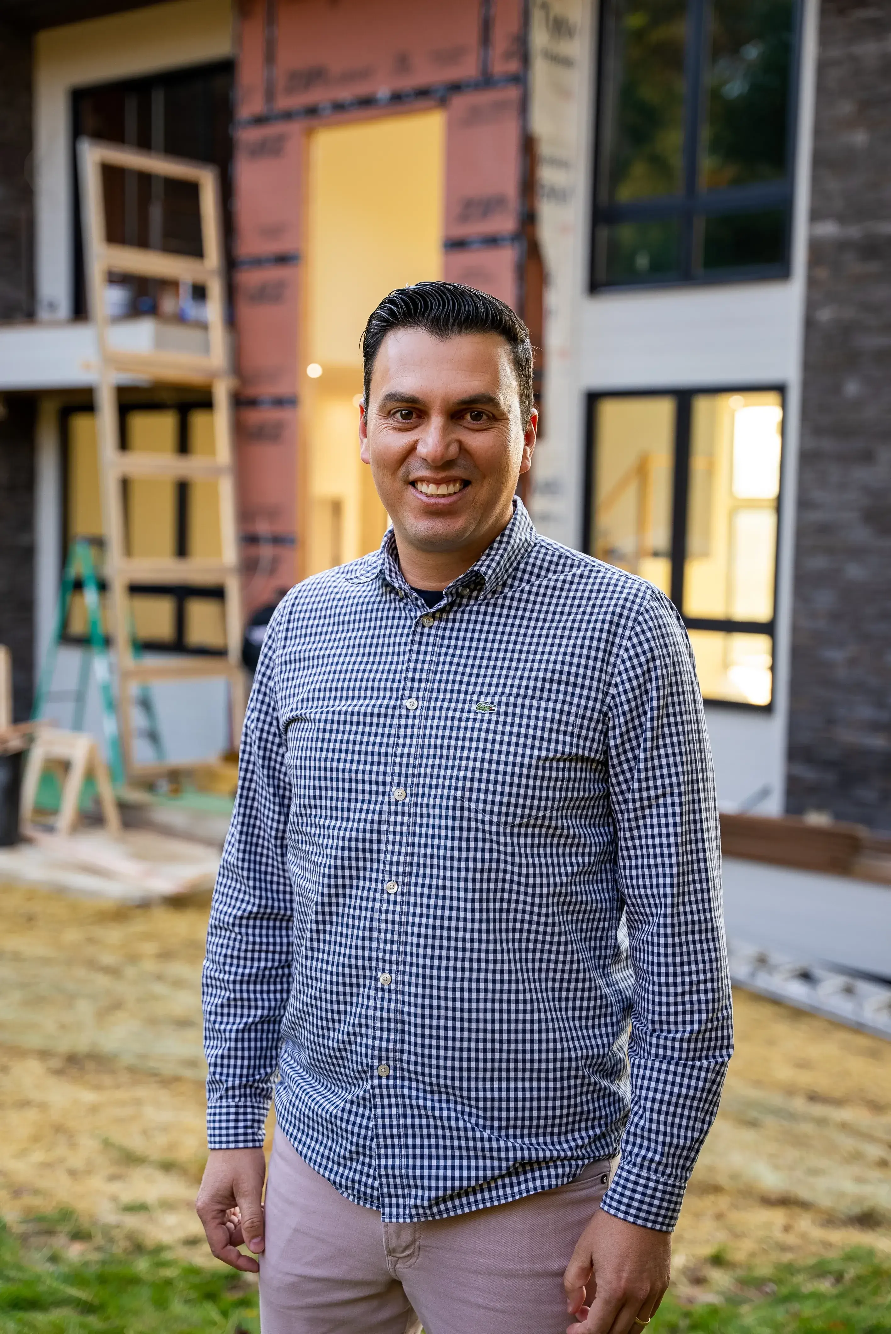 A smiling man in a checkered shirt standing in front of a construction site with an orange border accentuating the image.