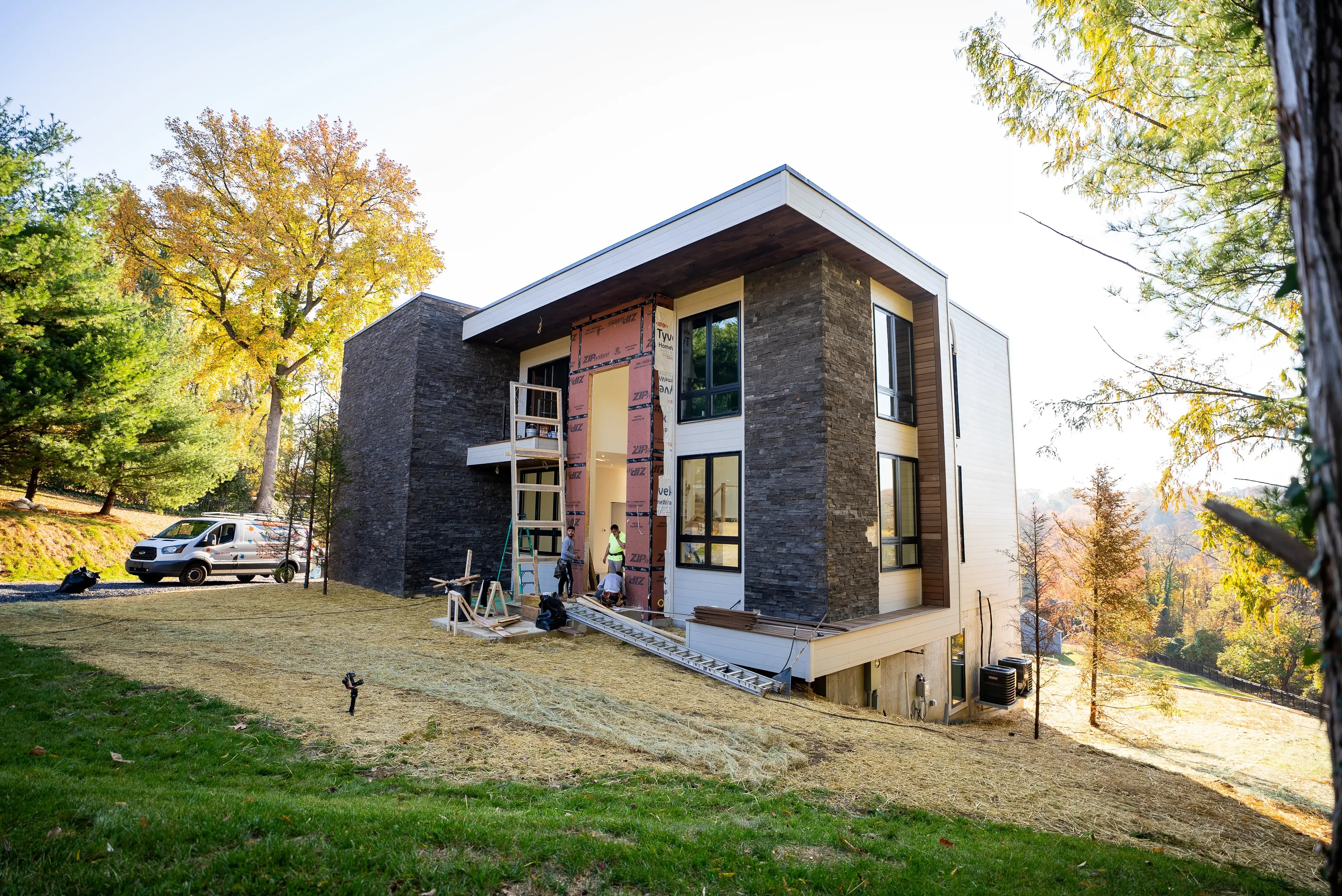 A modern house under construction, surrounded by lush greenery and autumn-colored trees, with workers actively completing the facade and entryway details.