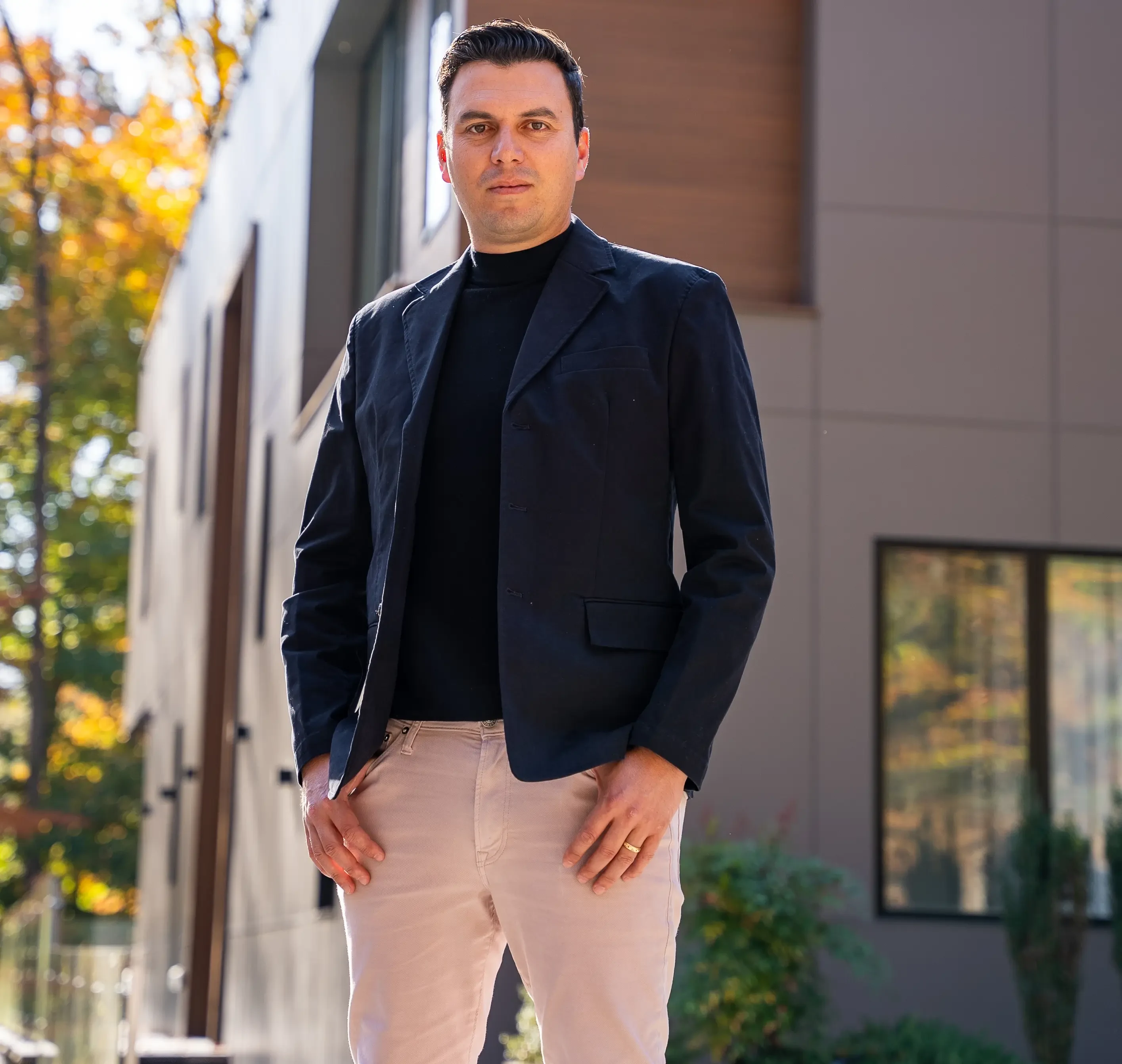 A confident man dressed in a dark blazer and light pants stands in front of a modern house, with autumnal trees in the background.