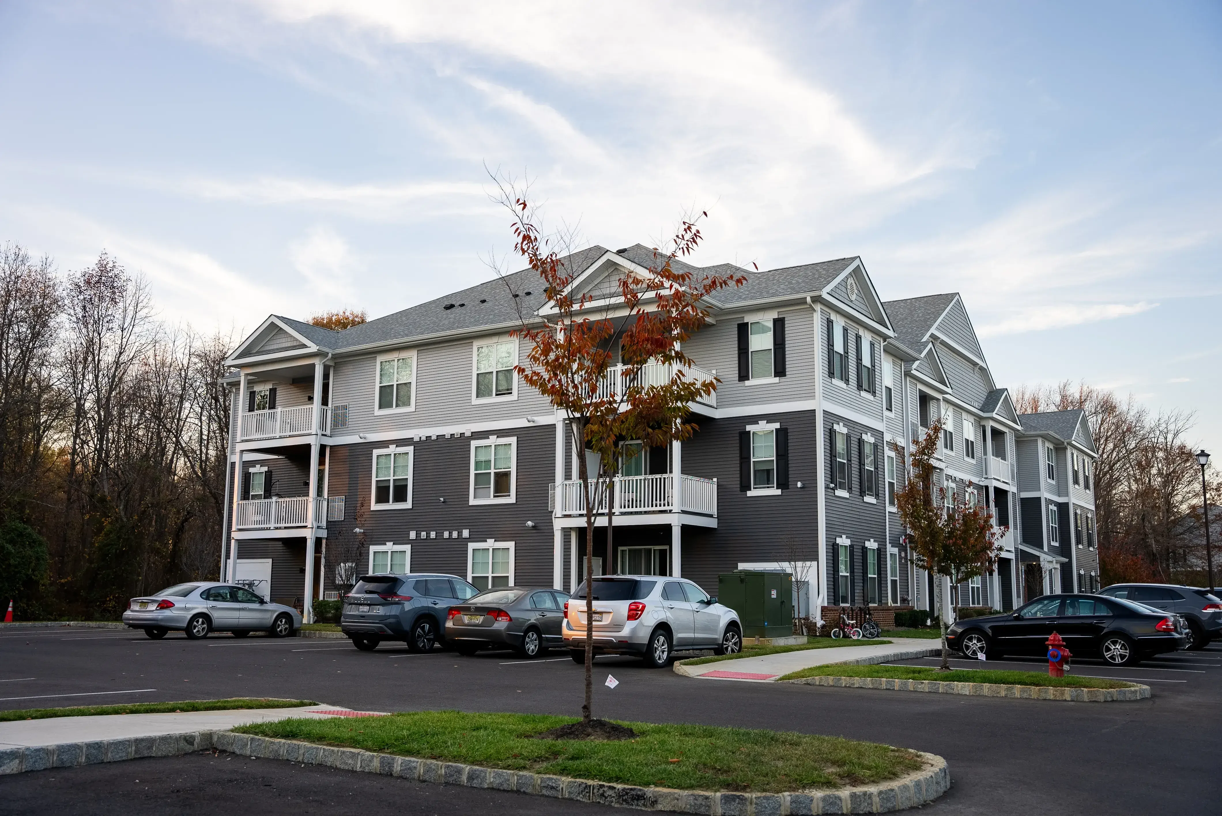 A modern apartment building with gray siding and white trim, surrounded by trees and parked cars in the foreground.