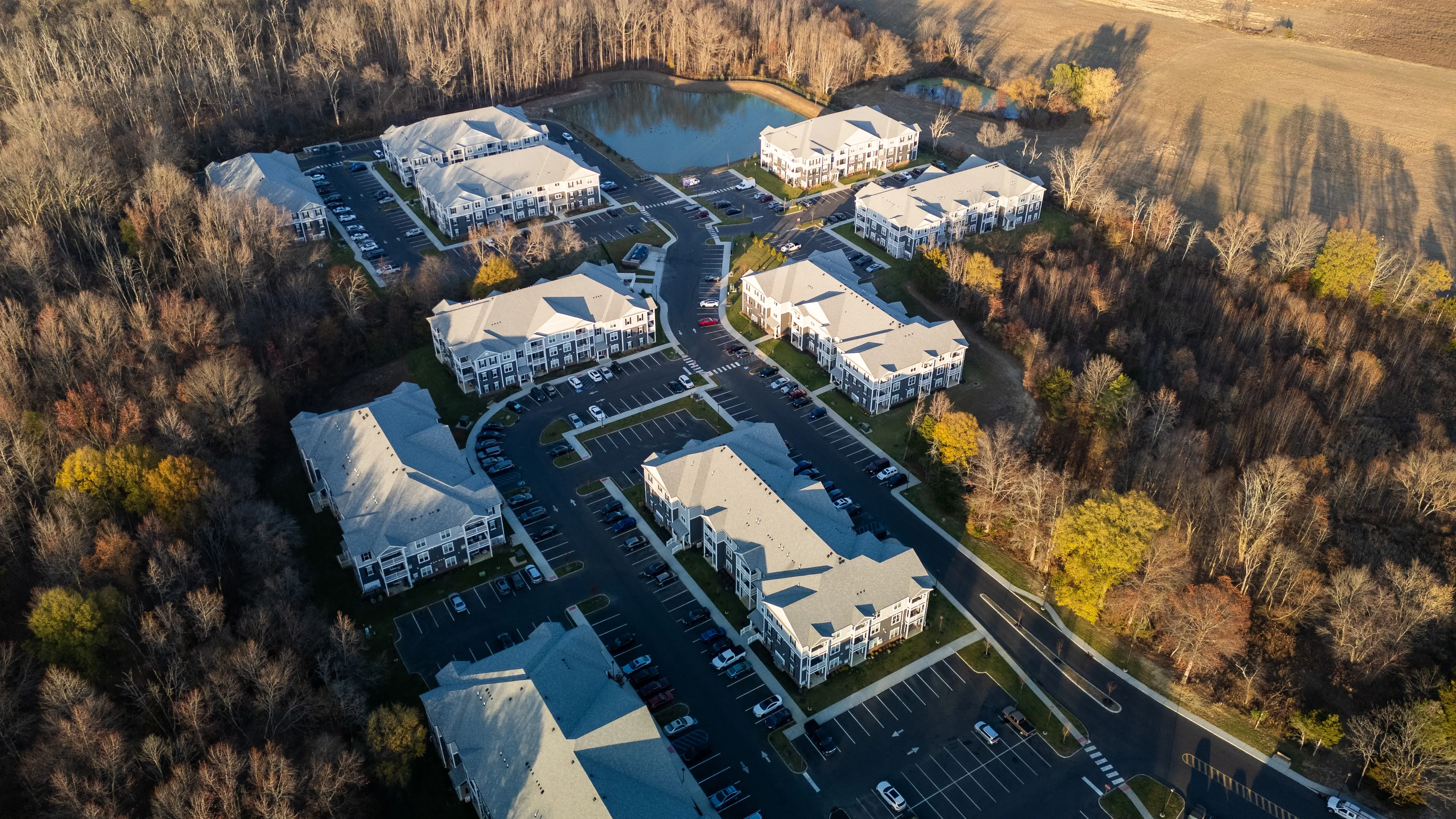 An aerial view of a modern apartment complex surrounded by dense autumnal woods and a serene pond, showcasing organized parking lots and well-maintained pathways connecting the buildings.